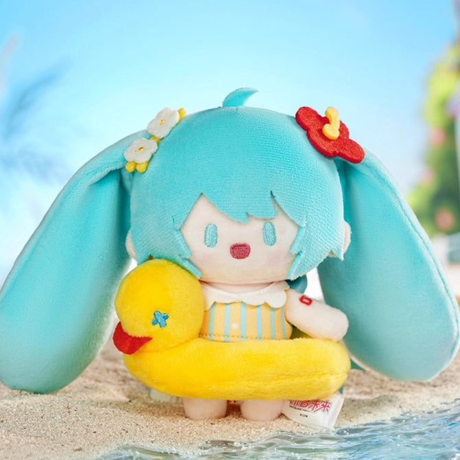 Hatsune Miku plush charm holding a yellow duck with striped swimsuit, on a sandy beach.