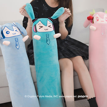 Long plush Miku pillows in blue, teal, and pink, held by a seated girl.