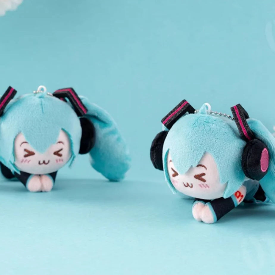 Two chibi-style Hatsune Miku plush charms lying down with smiling faces on a blue background