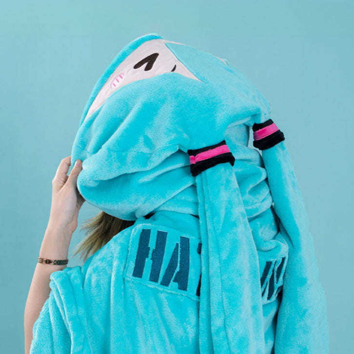 Hatsune Miku plush cape with "HATSUNE MIKU" embroidery, worn with the hood up, showing bunny ears